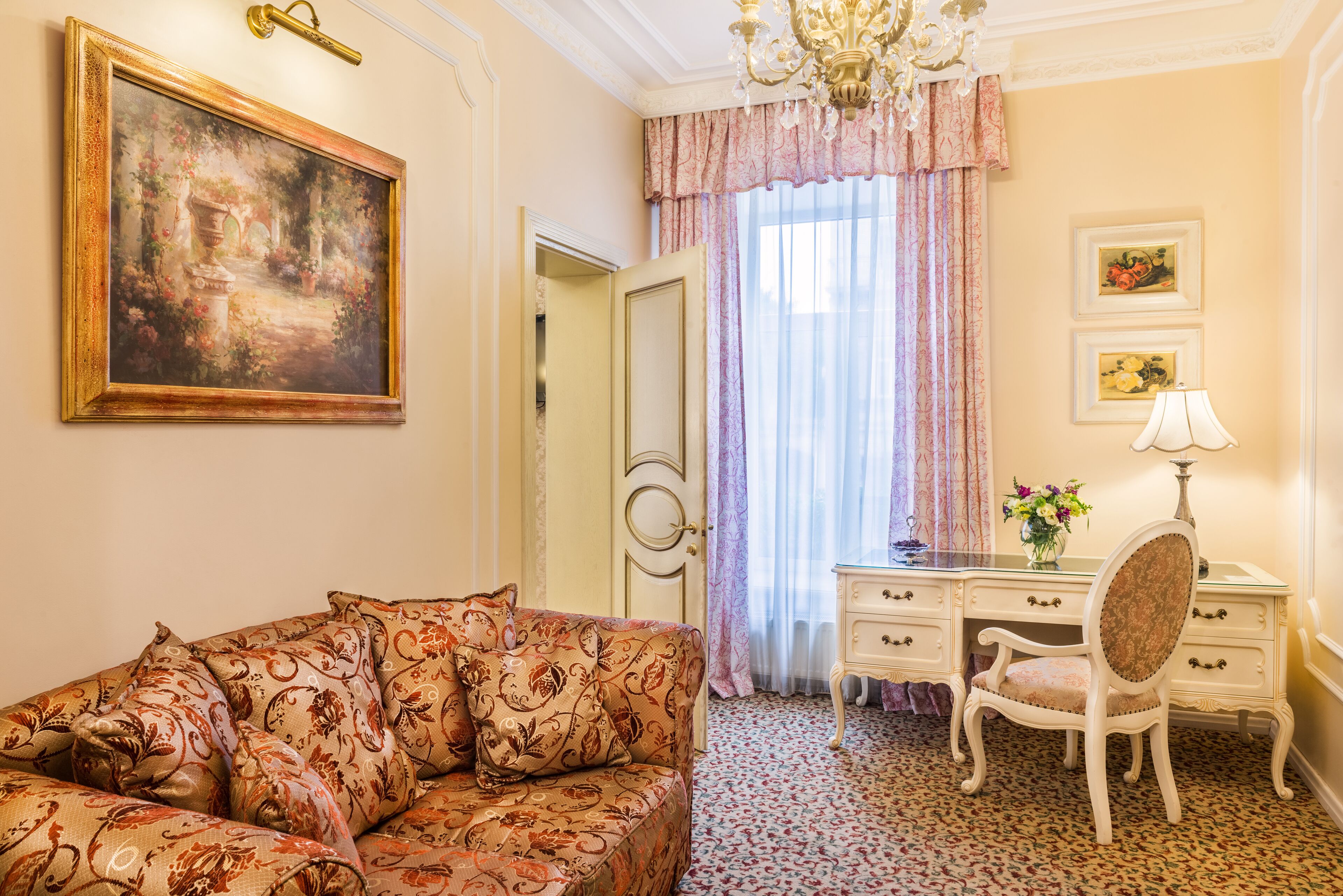 A room with a sofa, a desk, a chair, a chandelier, and a painting on the wall.
