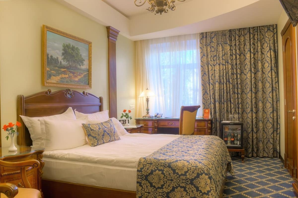 A hotel room with a large bed, a wooden headboard, a desk with a chair, a desk lamp, and a painting on the wall.