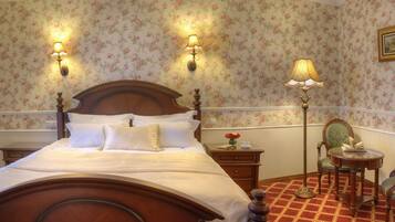 Un dormitorio clásico con una cama de madera, papel tapiz floral, dos lámparas fijadas a la pared, un sillón tapizado en verde y una mesita con un libro.