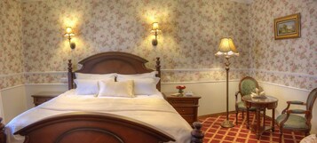 Un dormitorio clásico con una cama de madera, papel tapiz floral, dos lámparas fijadas a la pared, un sillón tapizado en verde y una mesita con un libro.