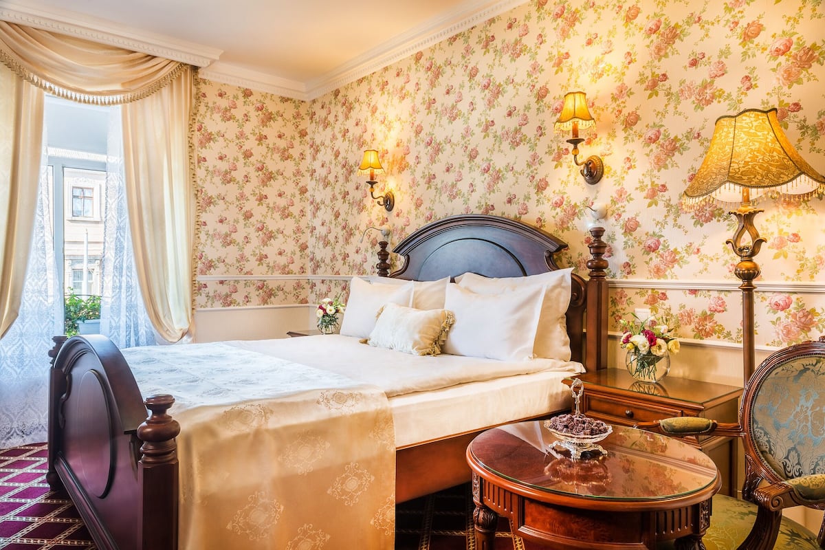 A bedroom with a large bed, floral wallpaper, two wall-mounted lamps, a wooden nightstand, and a small table with a vase of flowers.