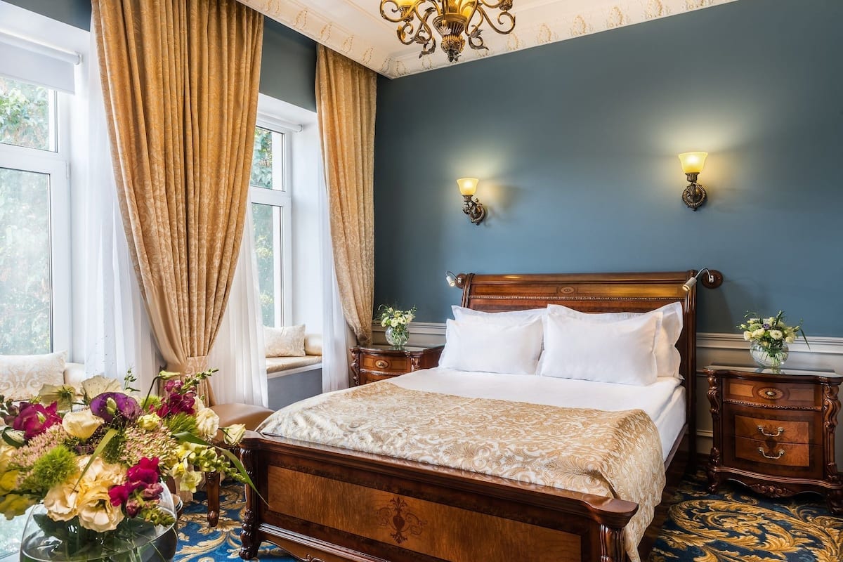 A bedroom with a wooden bed, two bedside tables, a nightstand, a vase with flowers, and a window with curtains.