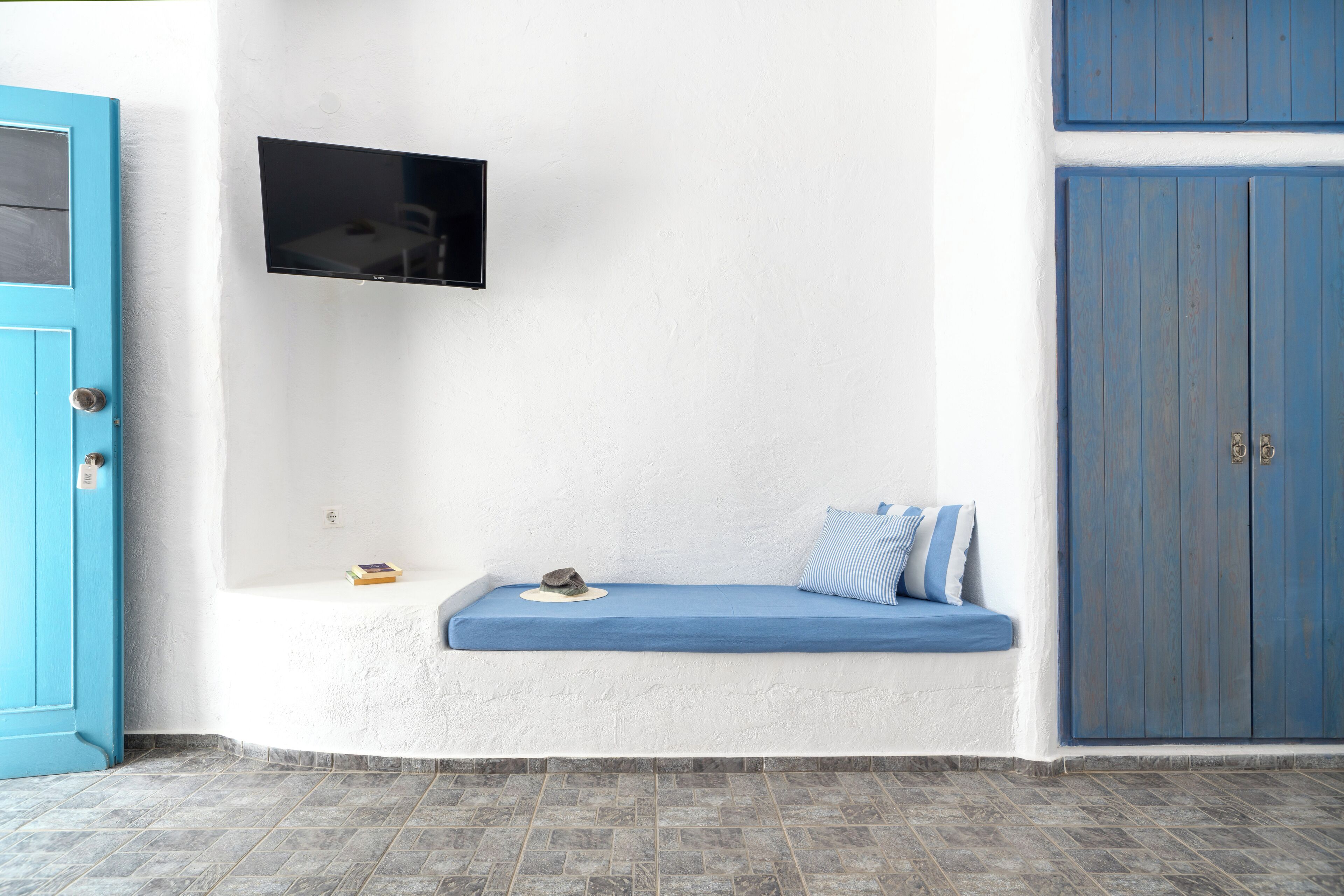 A room with a blue door, a mounted TV, a bench with cushions, and a wardrobe.