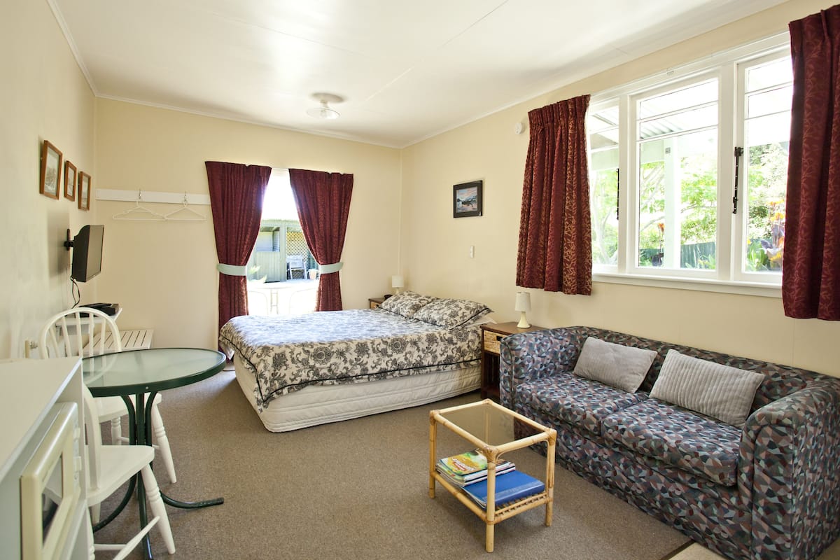 A room with a bed, a sofa, a small table with books, a TV, and a window with curtains.