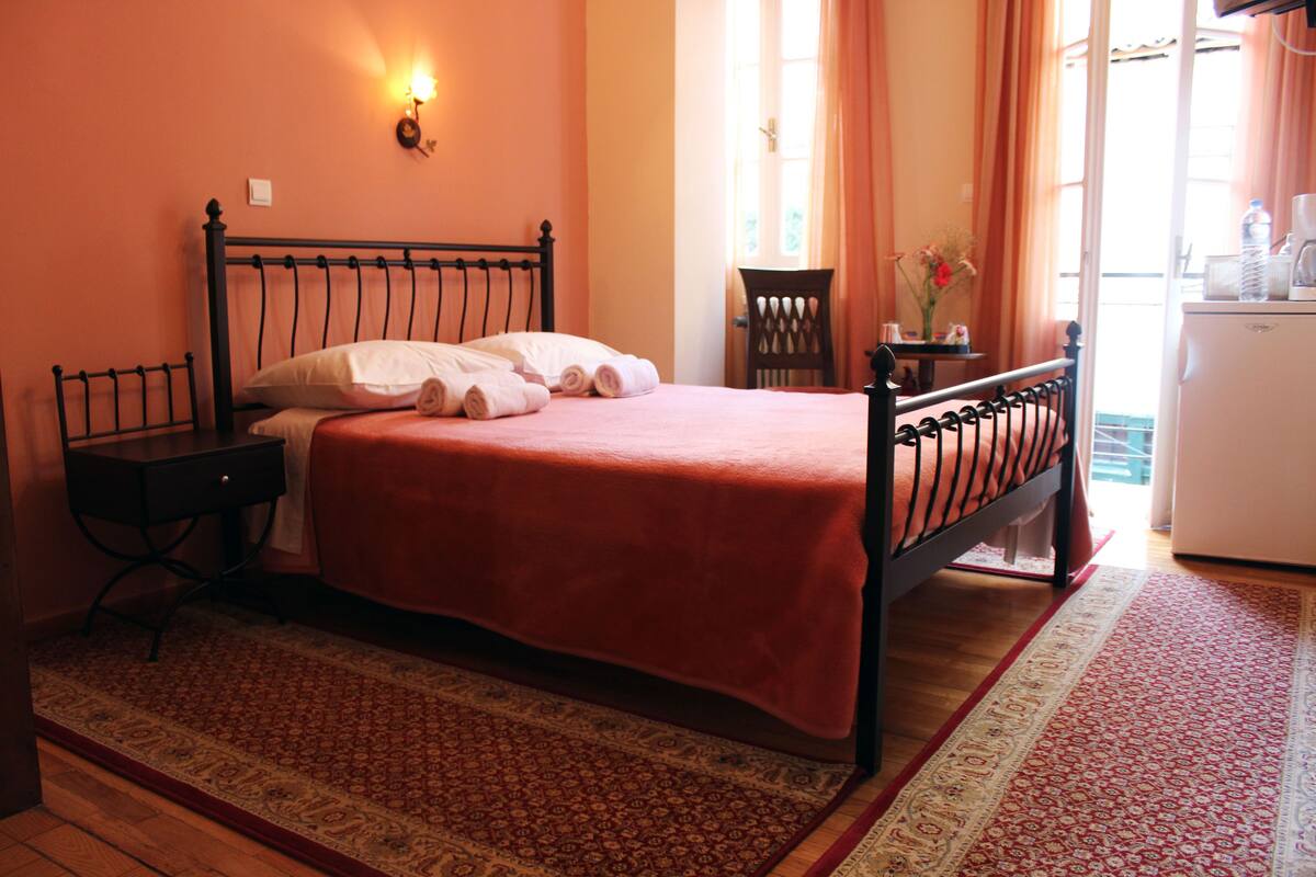 Una cama bien hecha con una colcha roja, una mesita de noche con una lámpara y una pequeña mesa con un jarrón de flores.