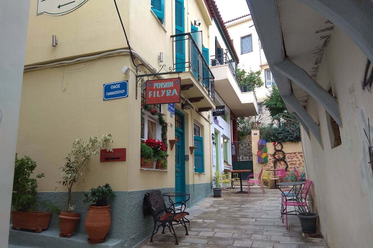 Una calle estrecha flanqueada por edificios, un letrero de recepción y mesas al aire libre con plantas en macetas.