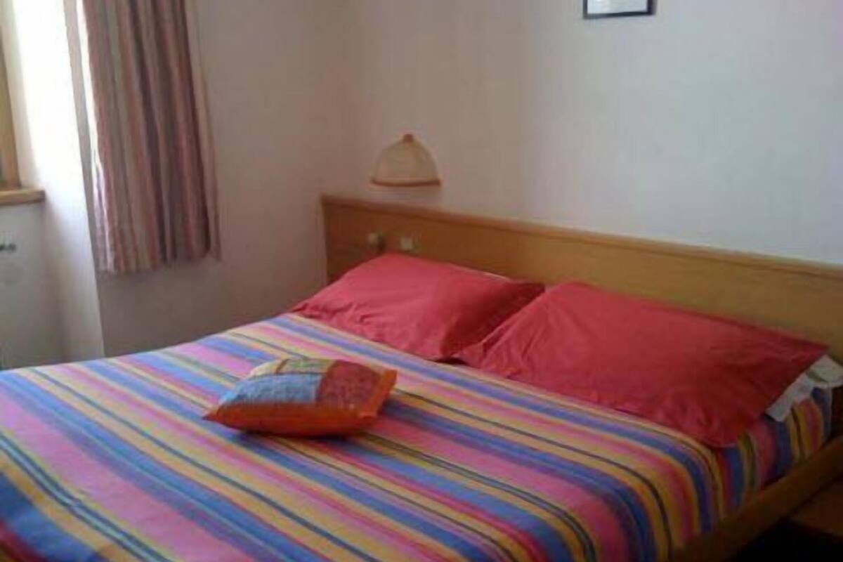Una cama con una colcha a rayas de colores y dos almohadas, una naranja y otra rosa, en una habitación con cabecera de madera y una lámpara.