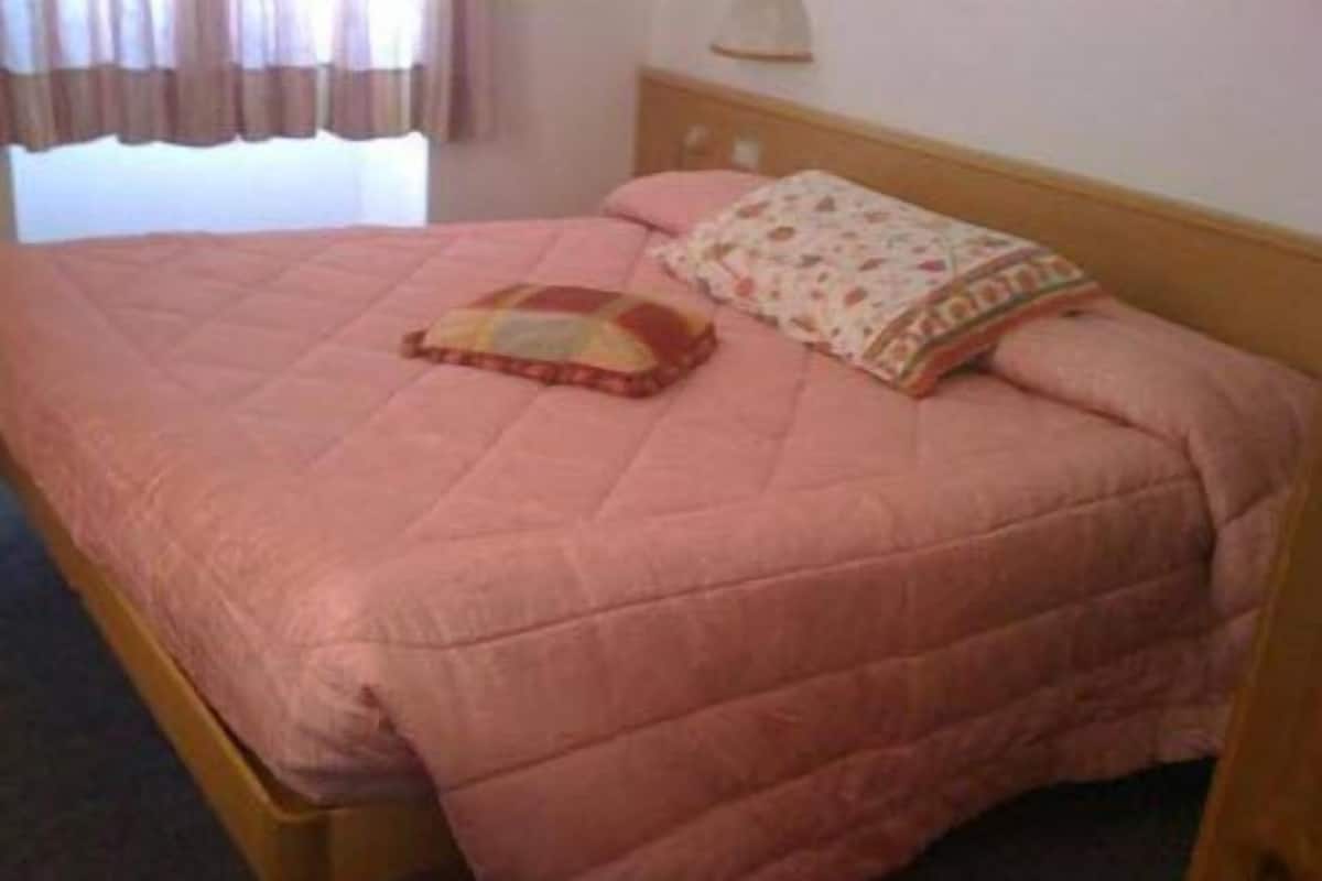 Una cama individual con colcha rosa y dos almohadas, cabecera de madera y una ventana con cortinas.