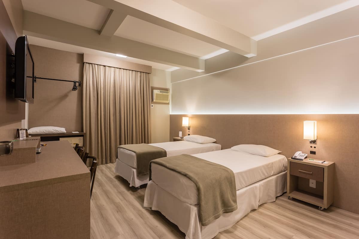 A hotel room with two beds, a desk, a flat-screen TV, a bedside table with a lamp, and an air conditioning unit.