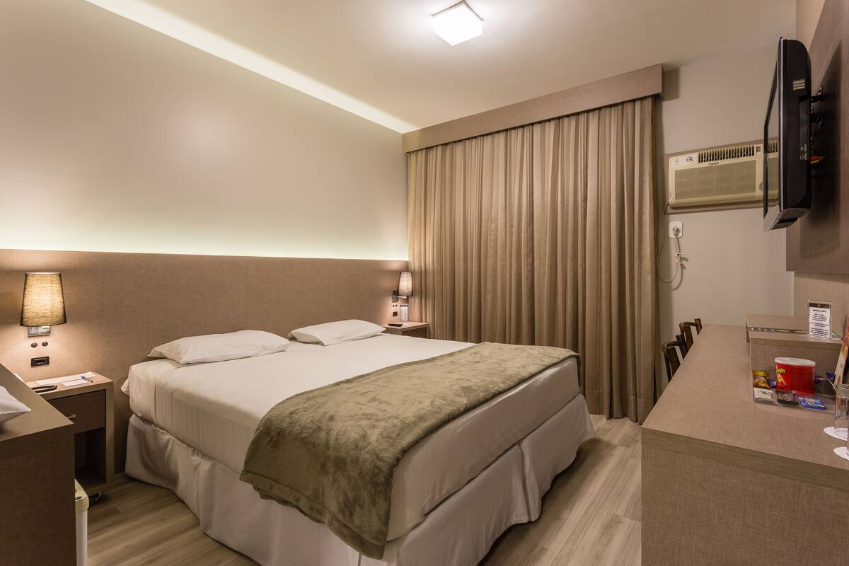 A hotel room with a bed, bedside tables, a desk, a TV, and a window with curtains.