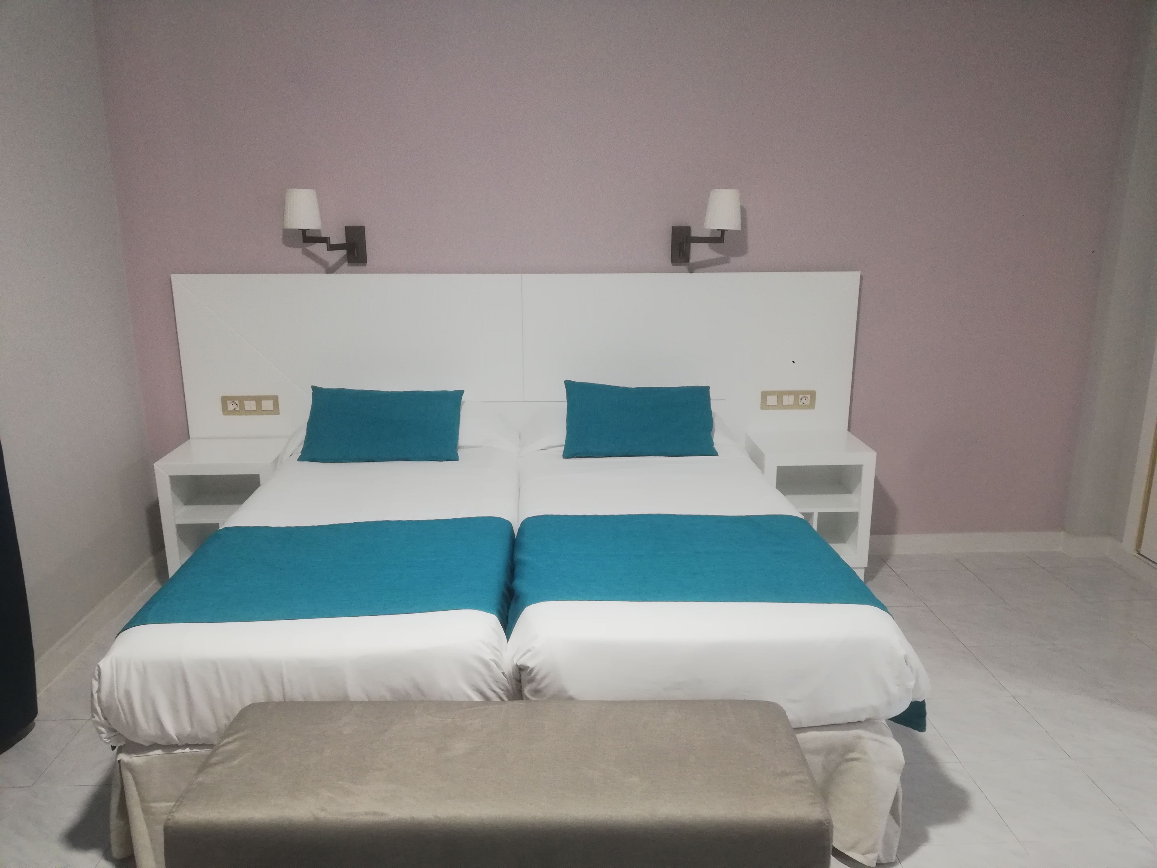 Ein Doppelbett mit weißer und türkisfarbener Bettwäsche, zwei Nachttische mit Lampen und eine Bank am Fußende des Bettes.
