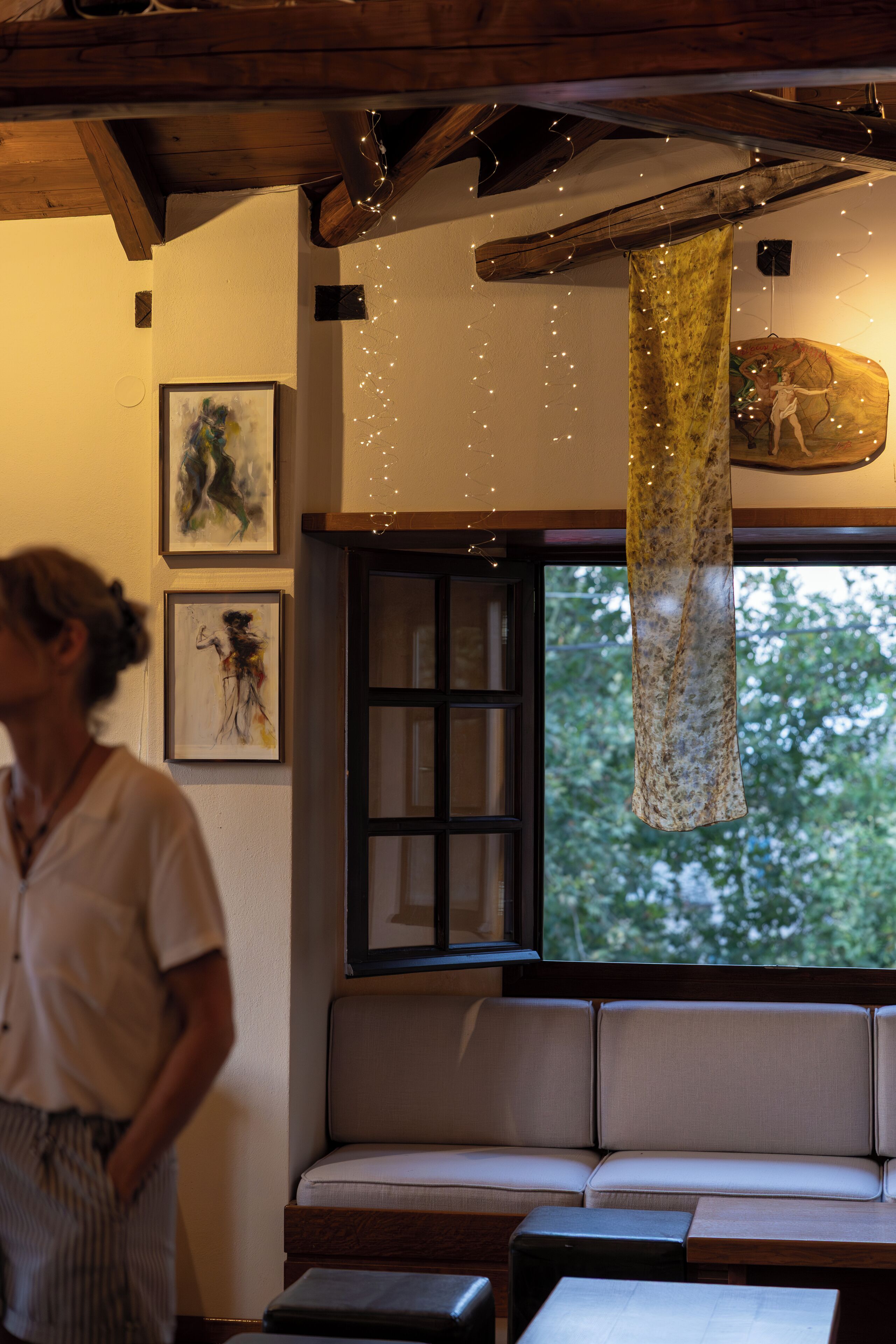 A room with a wooden ceiling, a bench with cushions, a window with a view of trees, and two framed artworks on the wall.