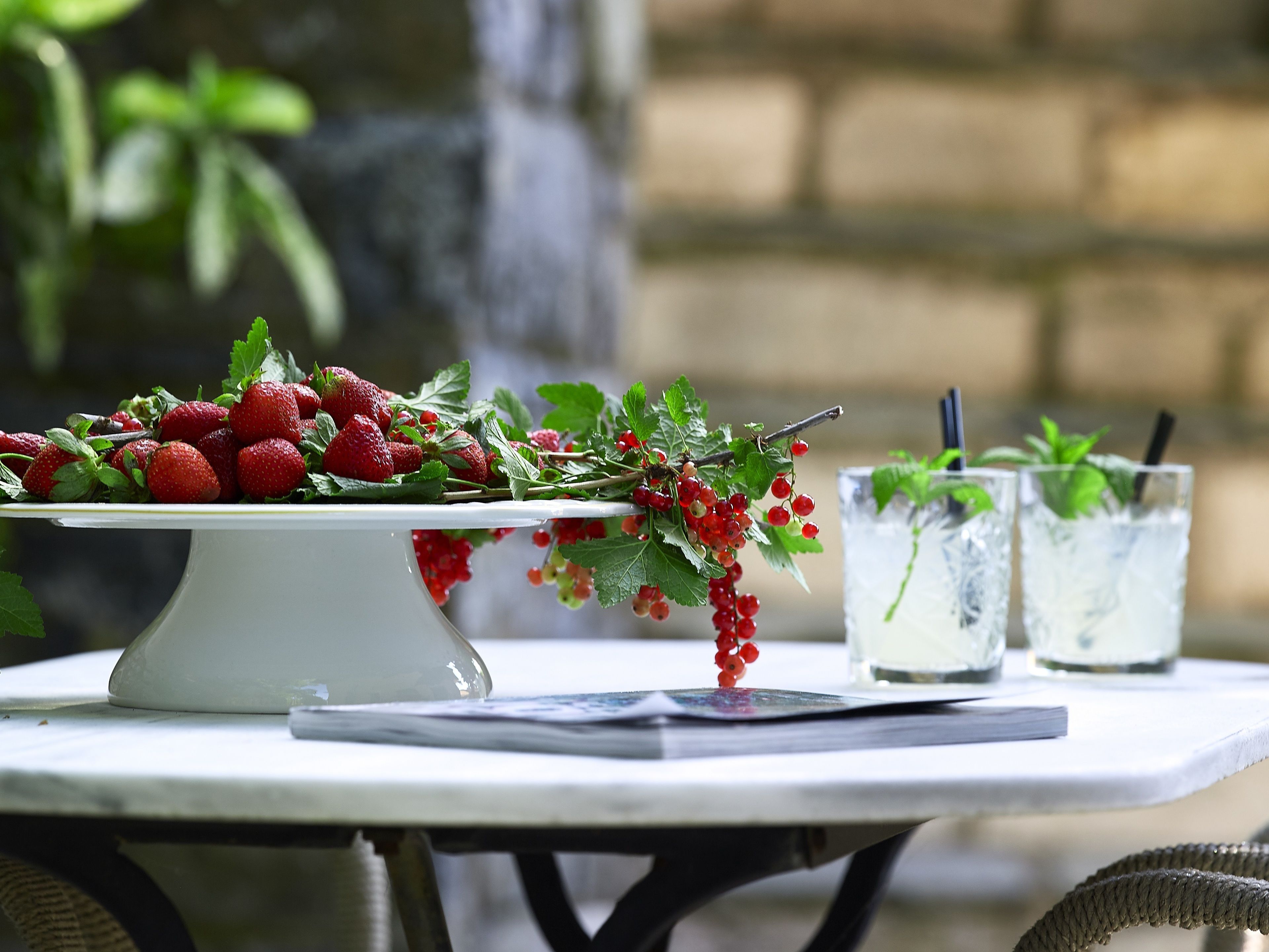 Una mesa con un recipiente de cerámica blanco que contiene fresas y bayas rojas, y dos vasos de bebida helada adornados con hojas de menta.