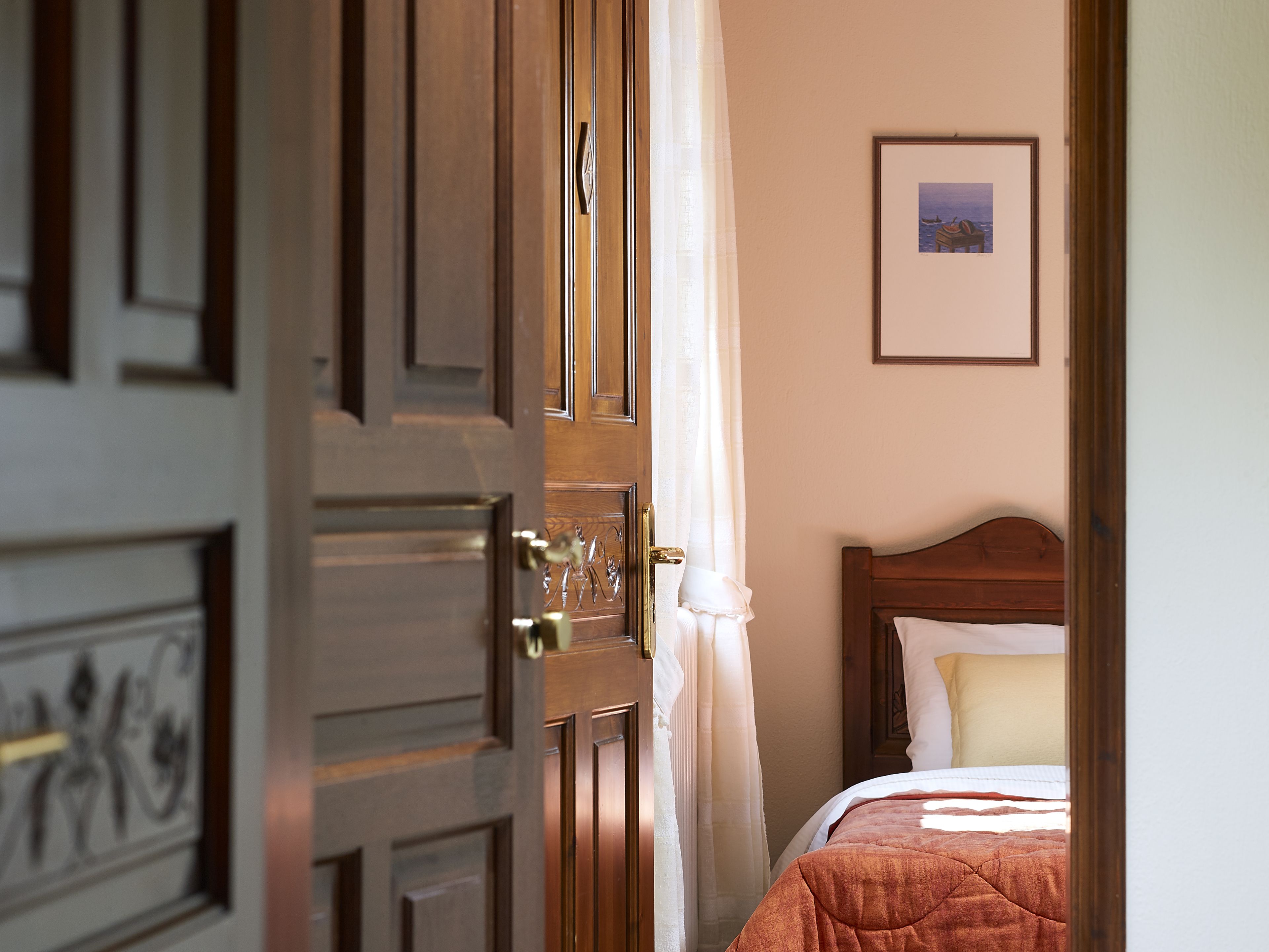 A hotel room with a wooden door, a bed with a quilt, a framed picture on the wall, and a window with curtains.