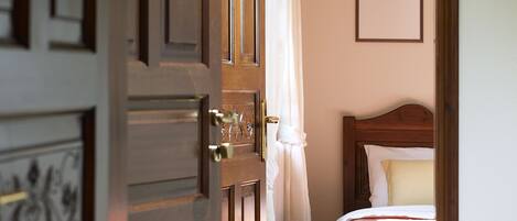 A hotel room with a wooden door, a bed with a quilt, a framed picture on the wall, and a window with curtains.
