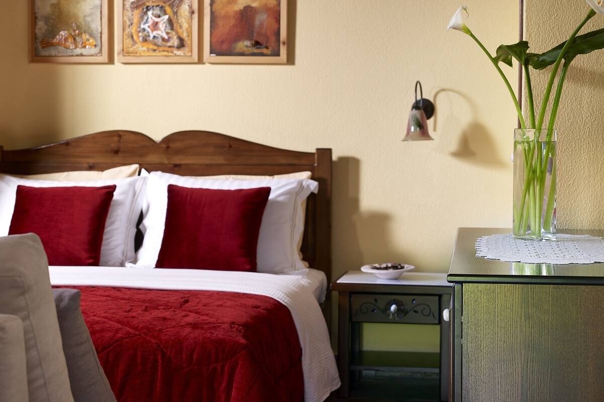 Una cama bien hecha con ropa de cama roja y blanca, una mesita de noche con un jarrón de flores y una pared con cuadros abstractos.