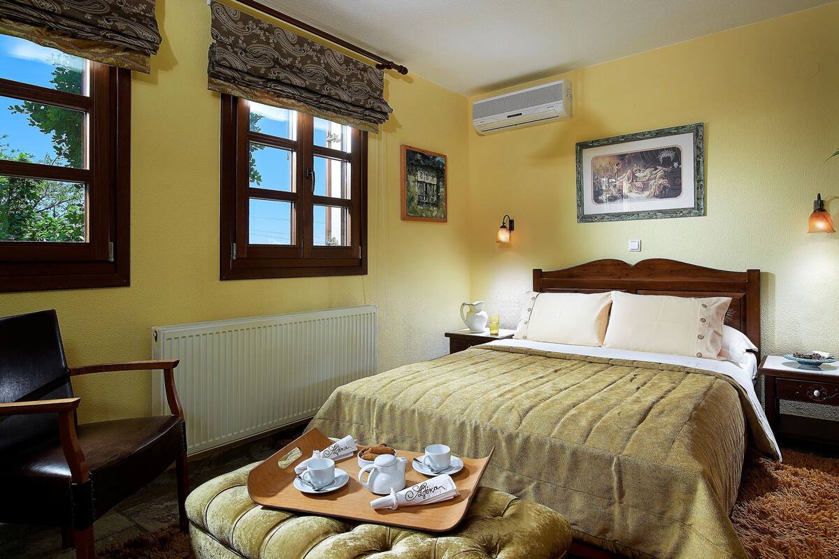 Un dormitorio con una cama, una silla de madera, una bandeja con juego de té, un radiador, una ventana con cortinas y un aire acondicionado fijado a la pared.