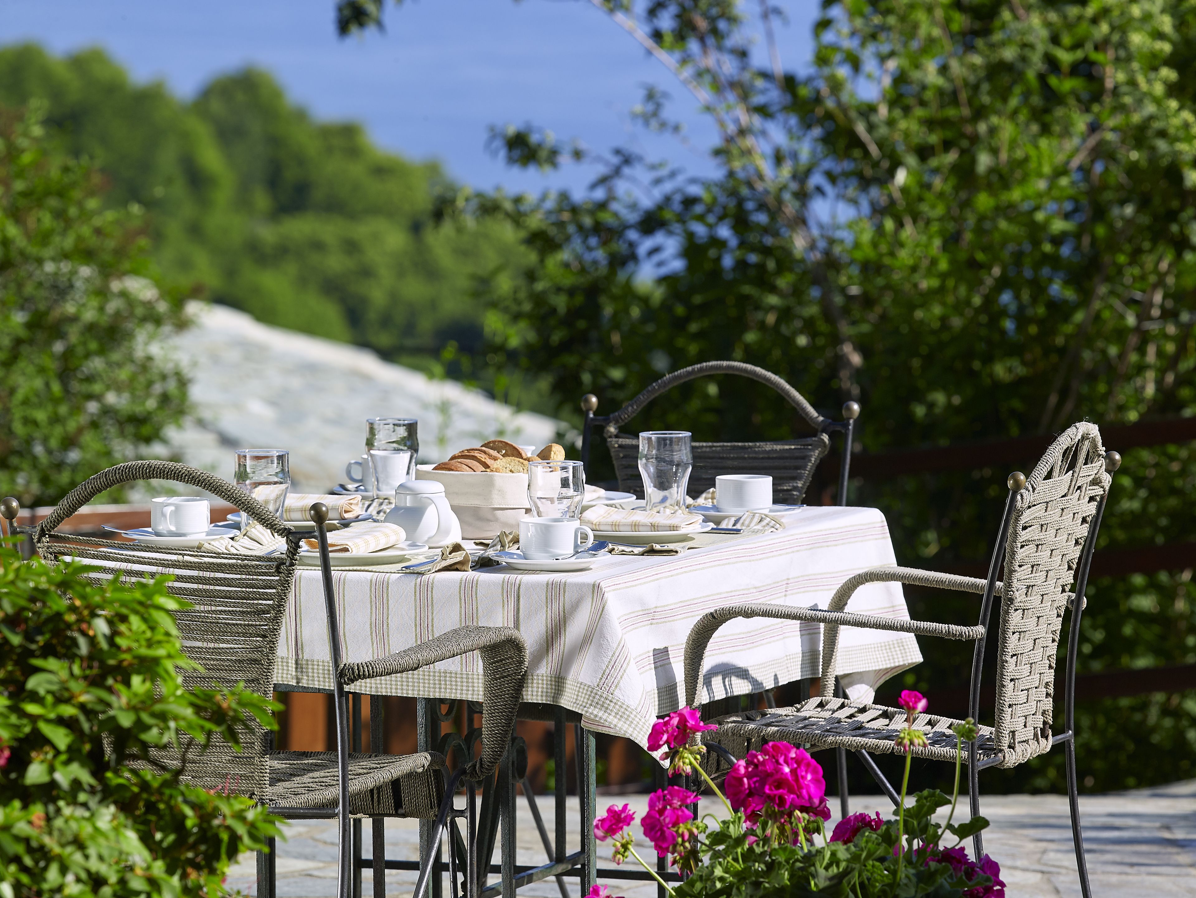 An outdoor dining table set with plates, cups, and a bread basket, surrounded by wicker chairs and pink flowers.