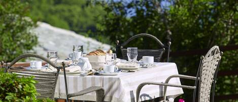 An outdoor dining table set with plates, cups, and a bread basket, surrounded by wicker chairs and pink flowers.