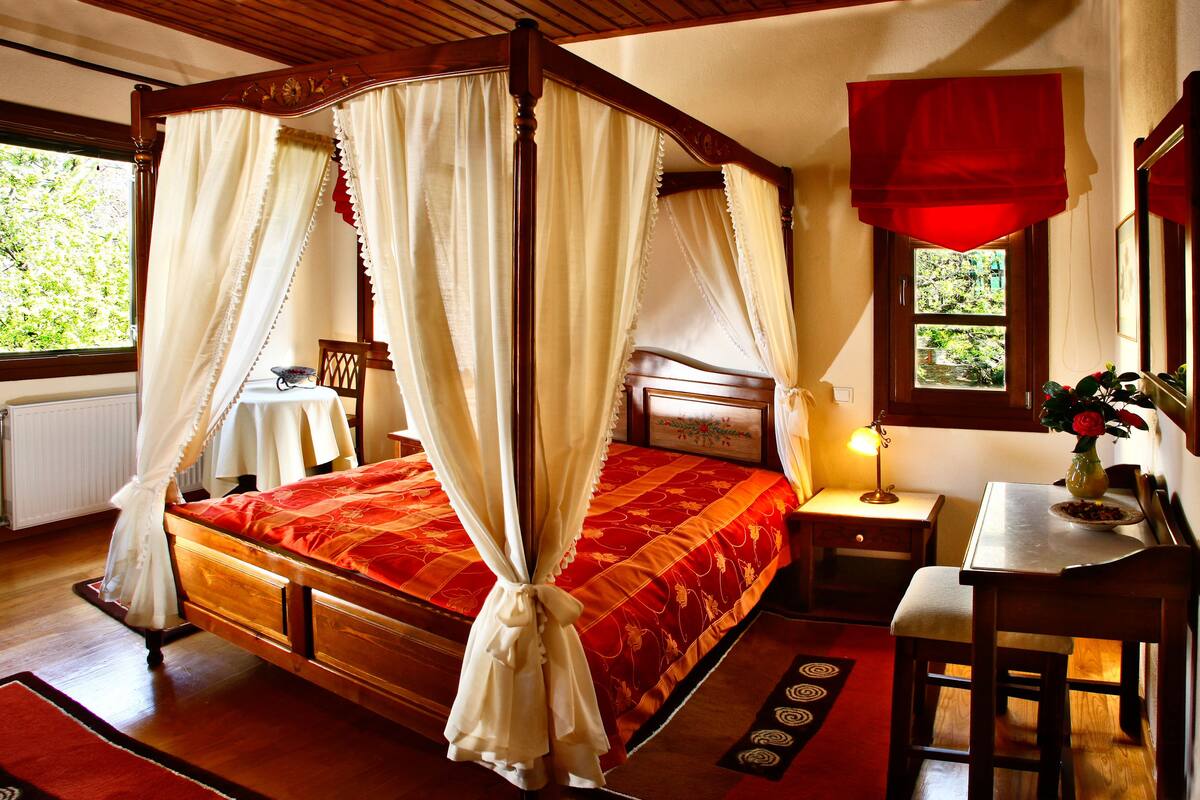 Una cama con dosel y cuatro columnas, con un edredón rojo, una mesita de noche con una lámpara y vistas al verdor exterior.