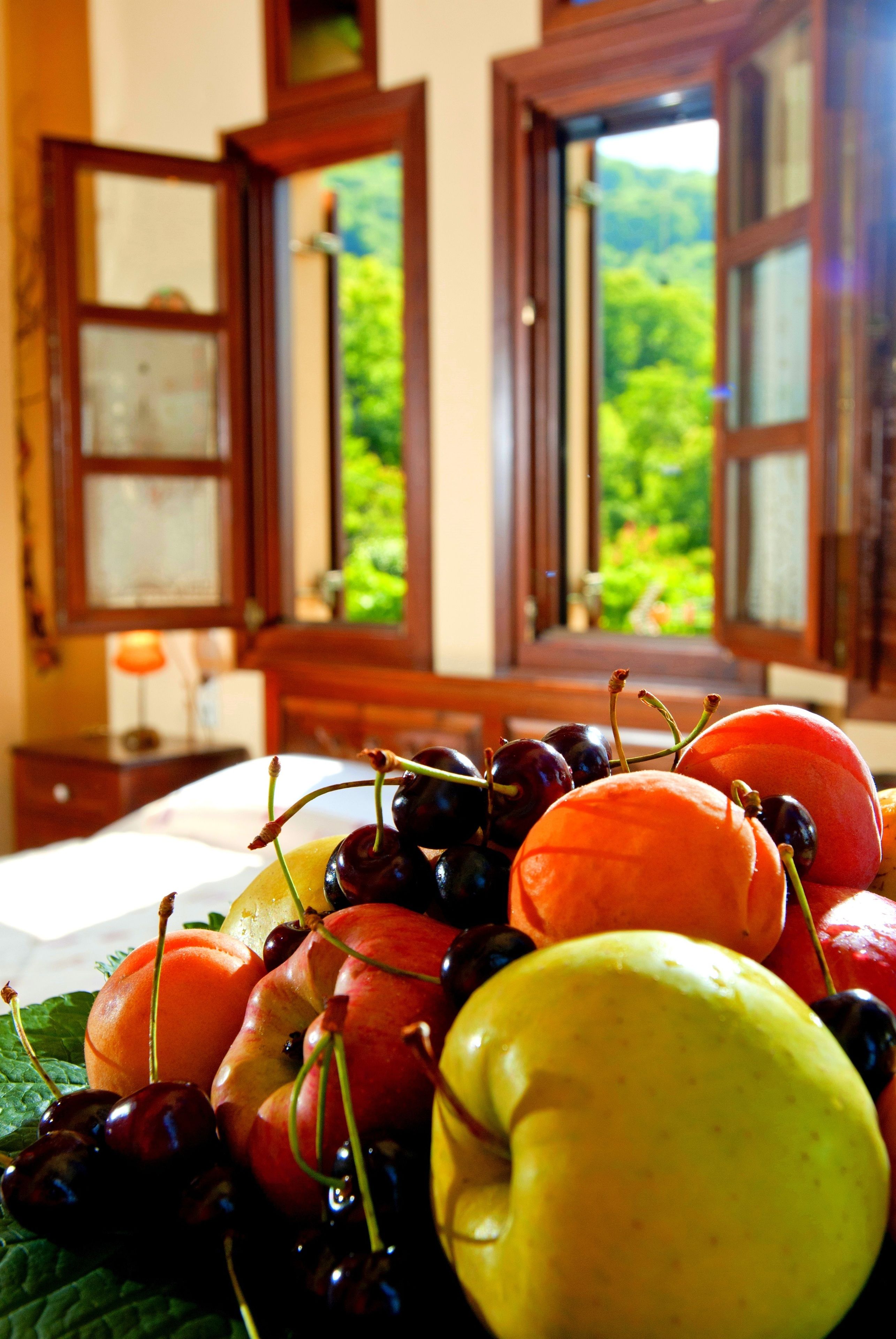 A table with a variety of fruits and a window with a view of greenery.