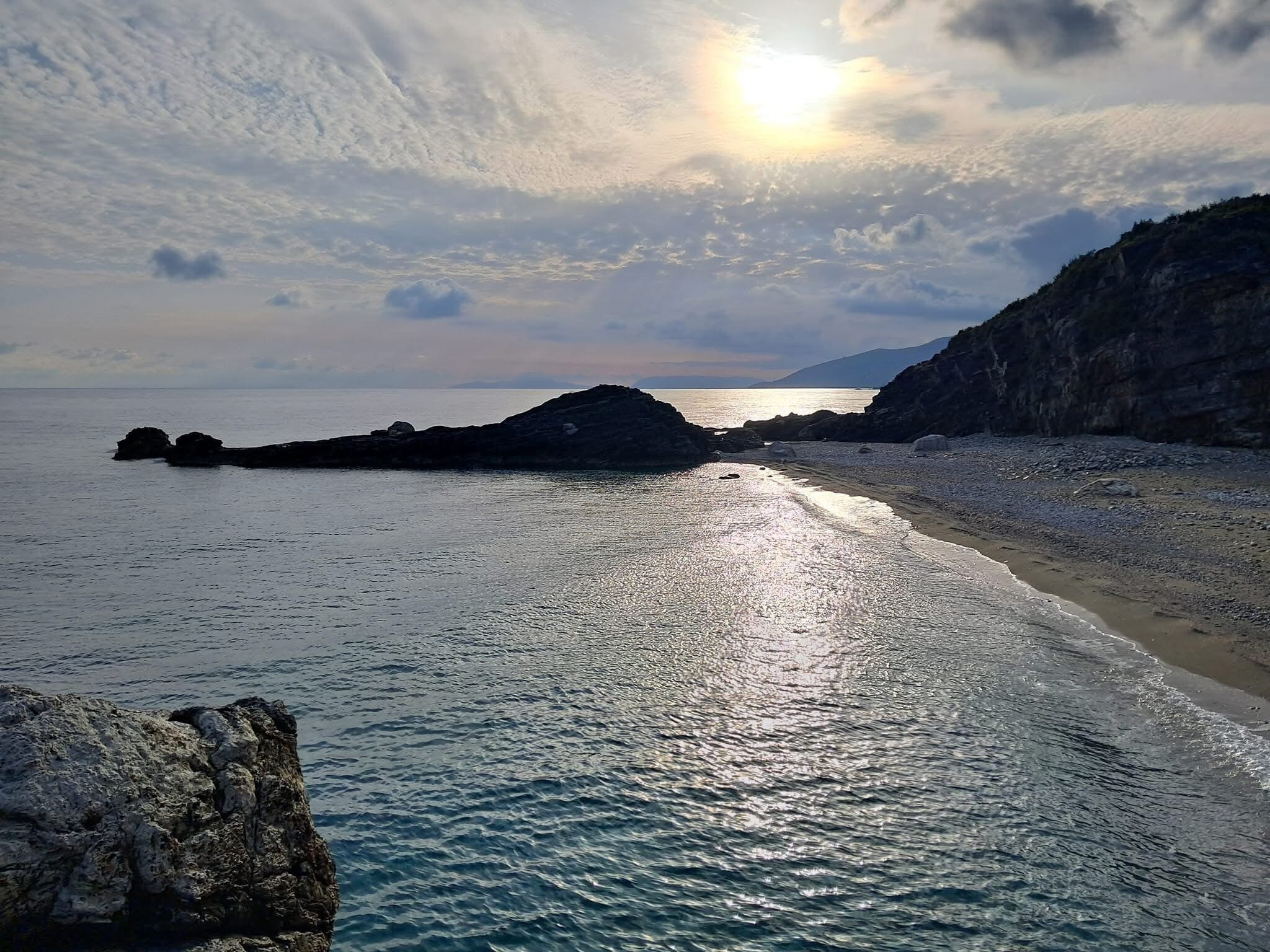 Escena costera con un litoral rocoso, mar tranquilo y una cadena montañosa distante bajo un cielo parcialmente nublado.