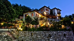 A two-story house with a stone wall in the foreground, a wooden staircase, and a garden with various plants and flowers. - Aleka's House (Zagora-Mouresi)