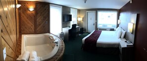 A hotel room with a large bathtub, a bed, a desk with a chair, a television, and a window with blinds.