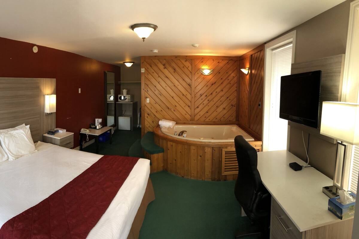 Une chambre d’hôtel avec un grand lit, un bureau, une chaise, une télévision, une lampe de bureau, un téléphone et une salle de bain avec une baignoire et un miroir.
