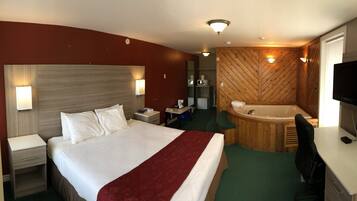 A hotel room with a large bed, a desk, a chair, a TV, a desk lamp, a telephone, and a bathroom with a bathtub and a mirror.