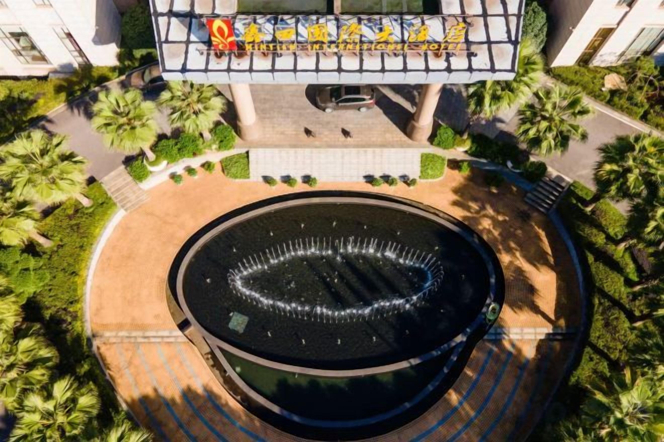 Una fuente circular rodeada por una zona pavimentada con palmeras, un edificio con un letrero y un cielo despejado.