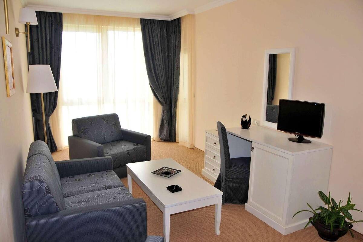 A hotel room with a sofa, a small table, a desk with a computer monitor, a mirror, and a potted plant.