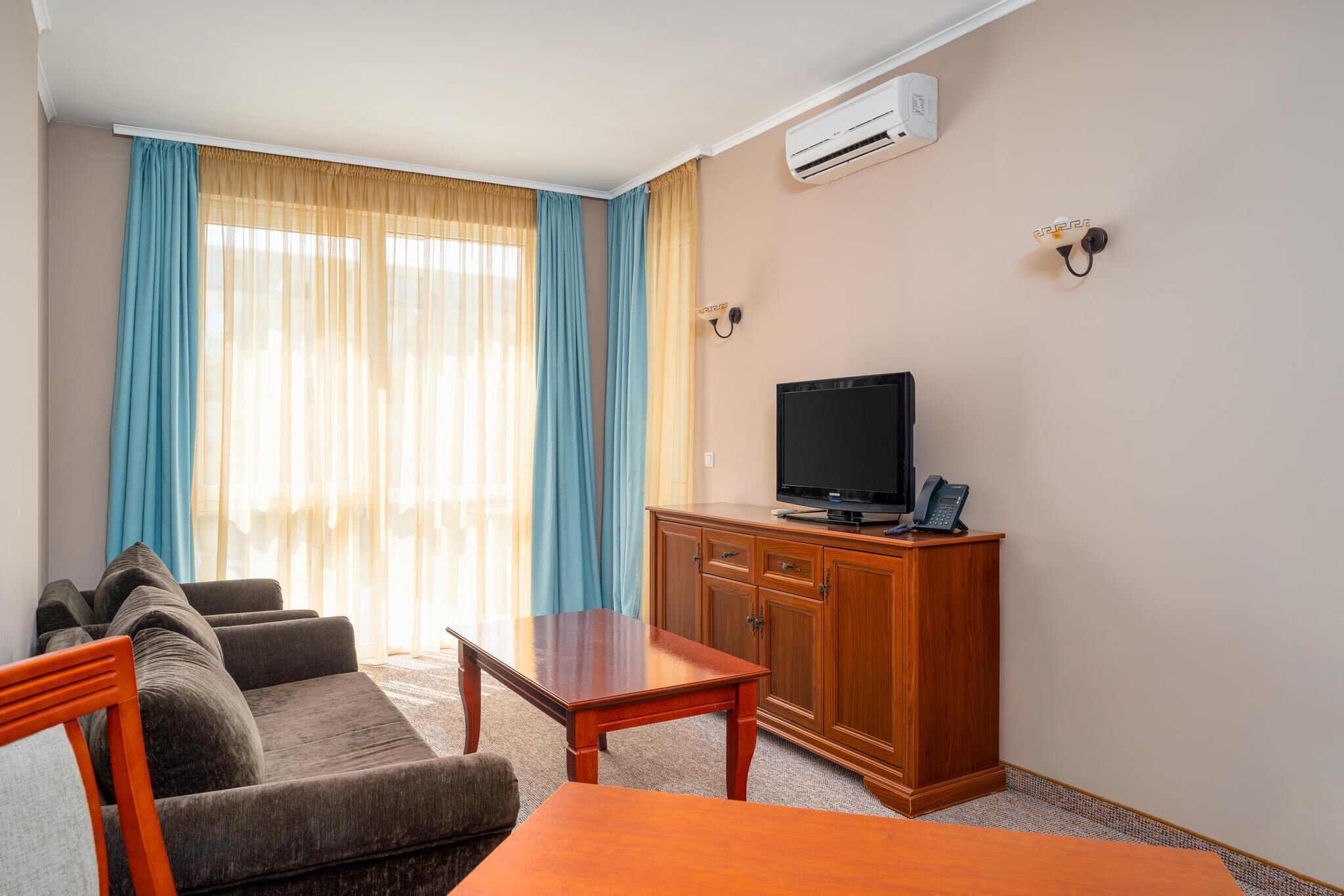 A hotel room with a sofa, a wooden table, a television on a cabinet, and an air conditioning unit on the wall.