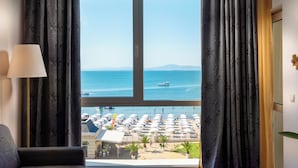 Een kamer met een groot raam met uitzicht op een strand met parasols en mensen.