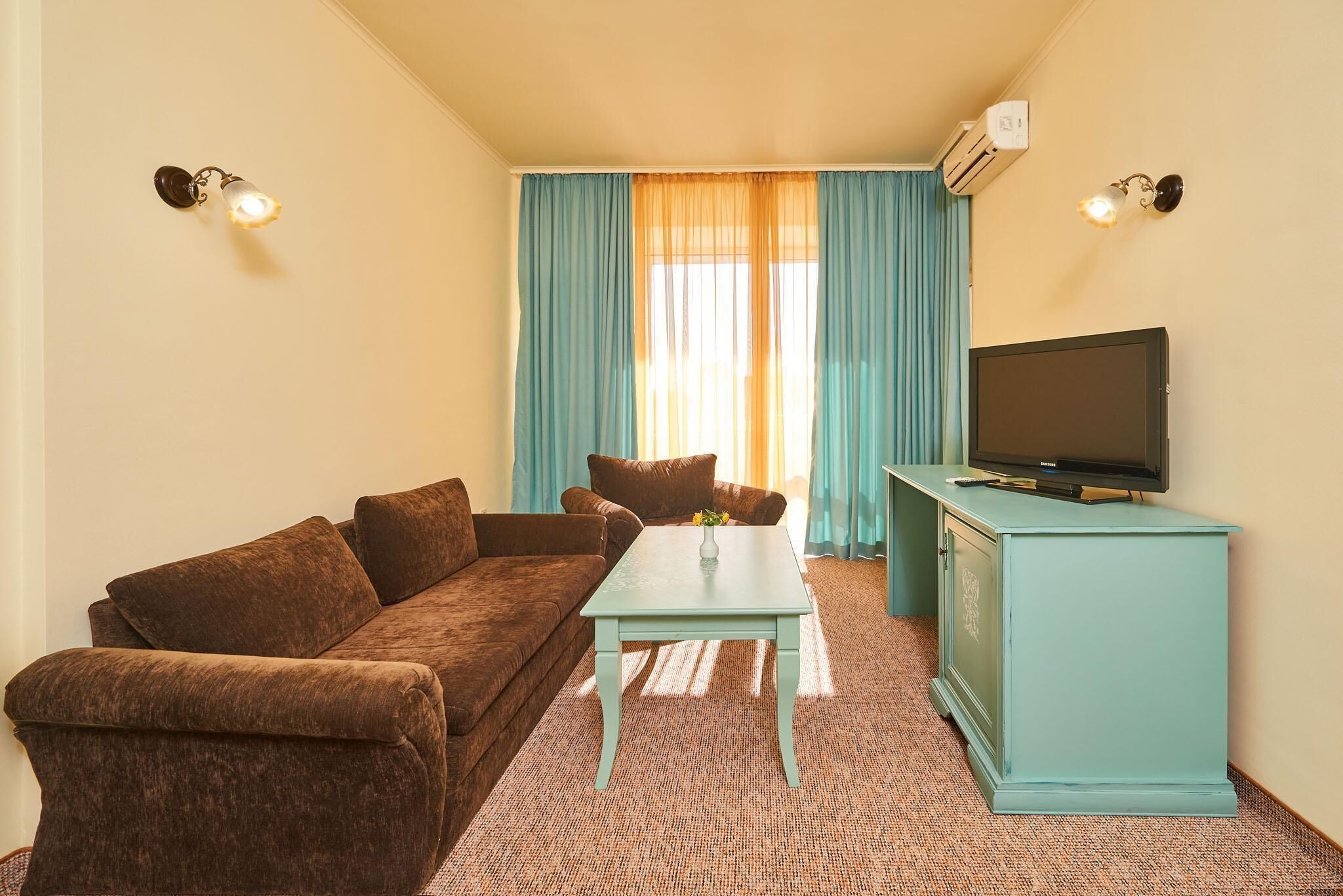 A living room with a brown sofa, a small table, a television on a cabinet, and a window with curtains.