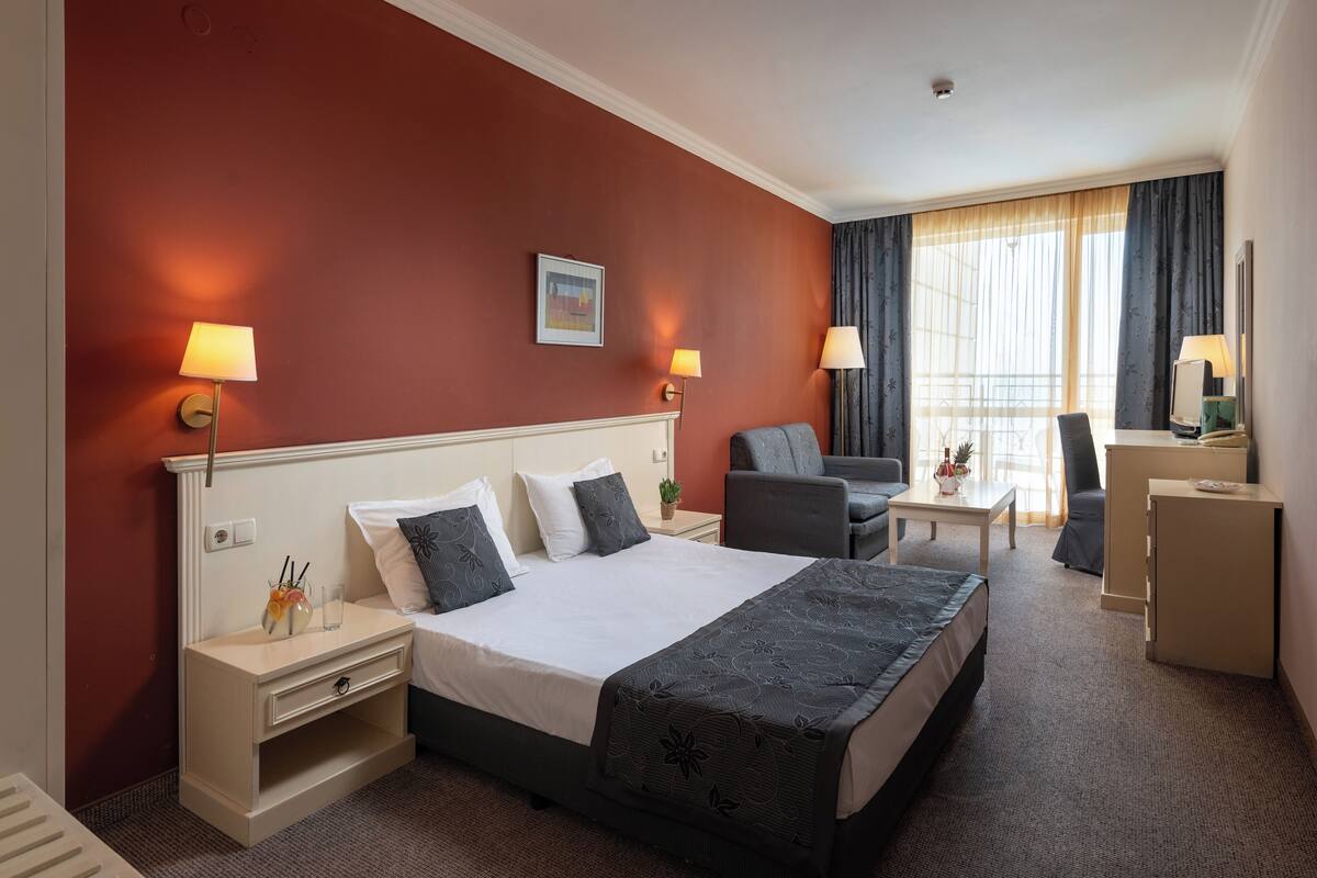 A hotel room with a large bed, a desk, a chair, a small table, and a nightstand.