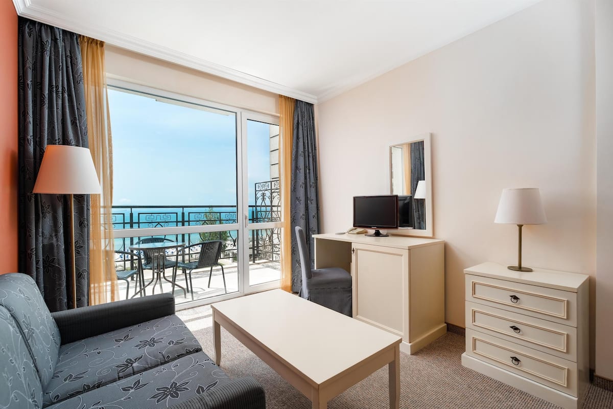 A hotel room with a sofa, a small table, a TV on a cabinet, and a balcony with outdoor seating.