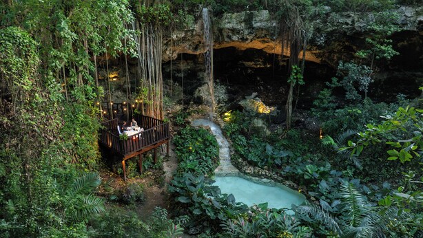 A wooden deck with a table and chairs overlooking a natural pool and waterfall.