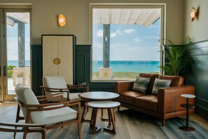A modern living room with a view of the ocean, featuring a brown leather sofa, white armchairs, and a round coffee table.