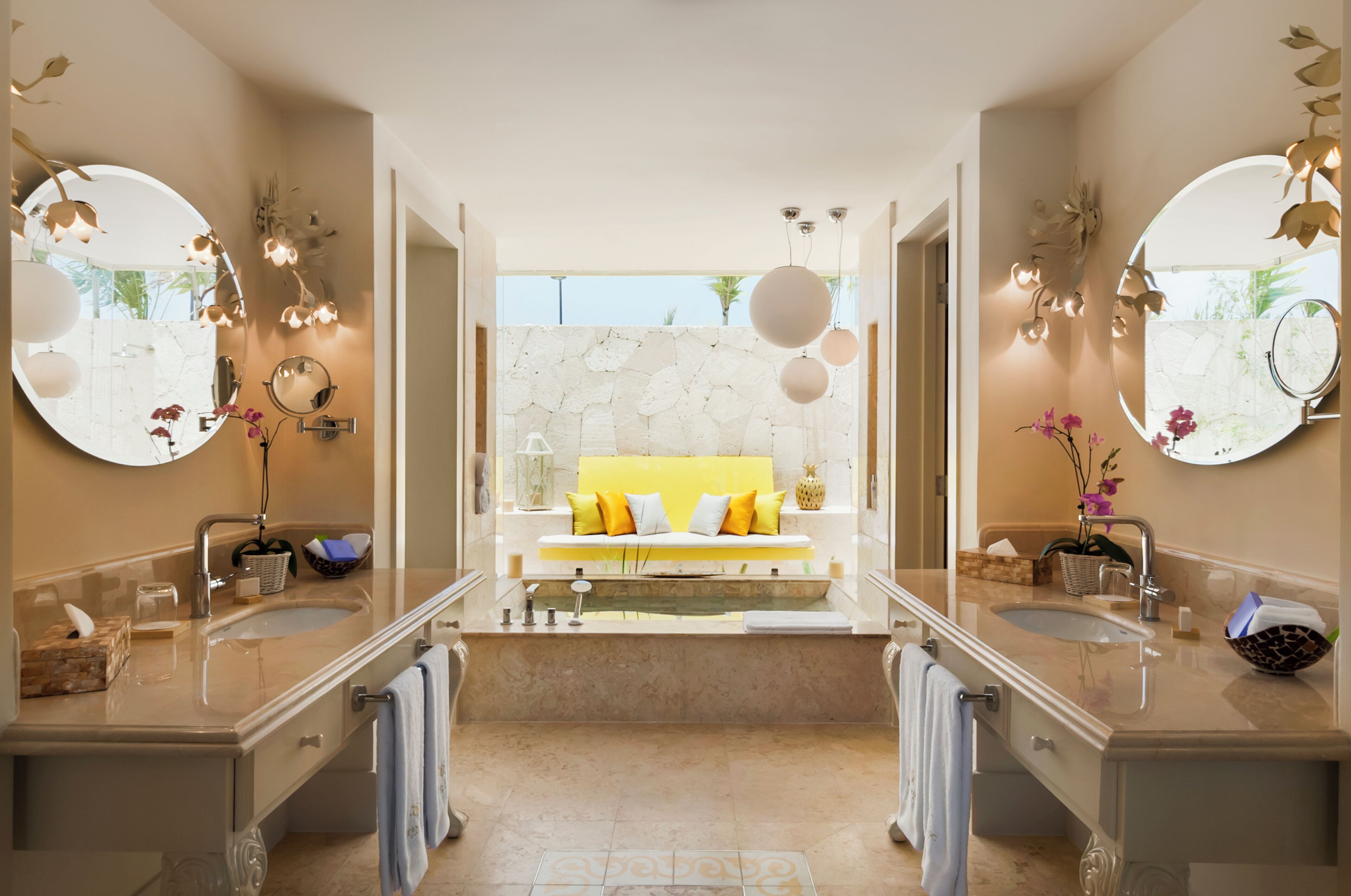 A modern bathroom with two sinks, a large mirror, a yellow bench, and a stone wall.