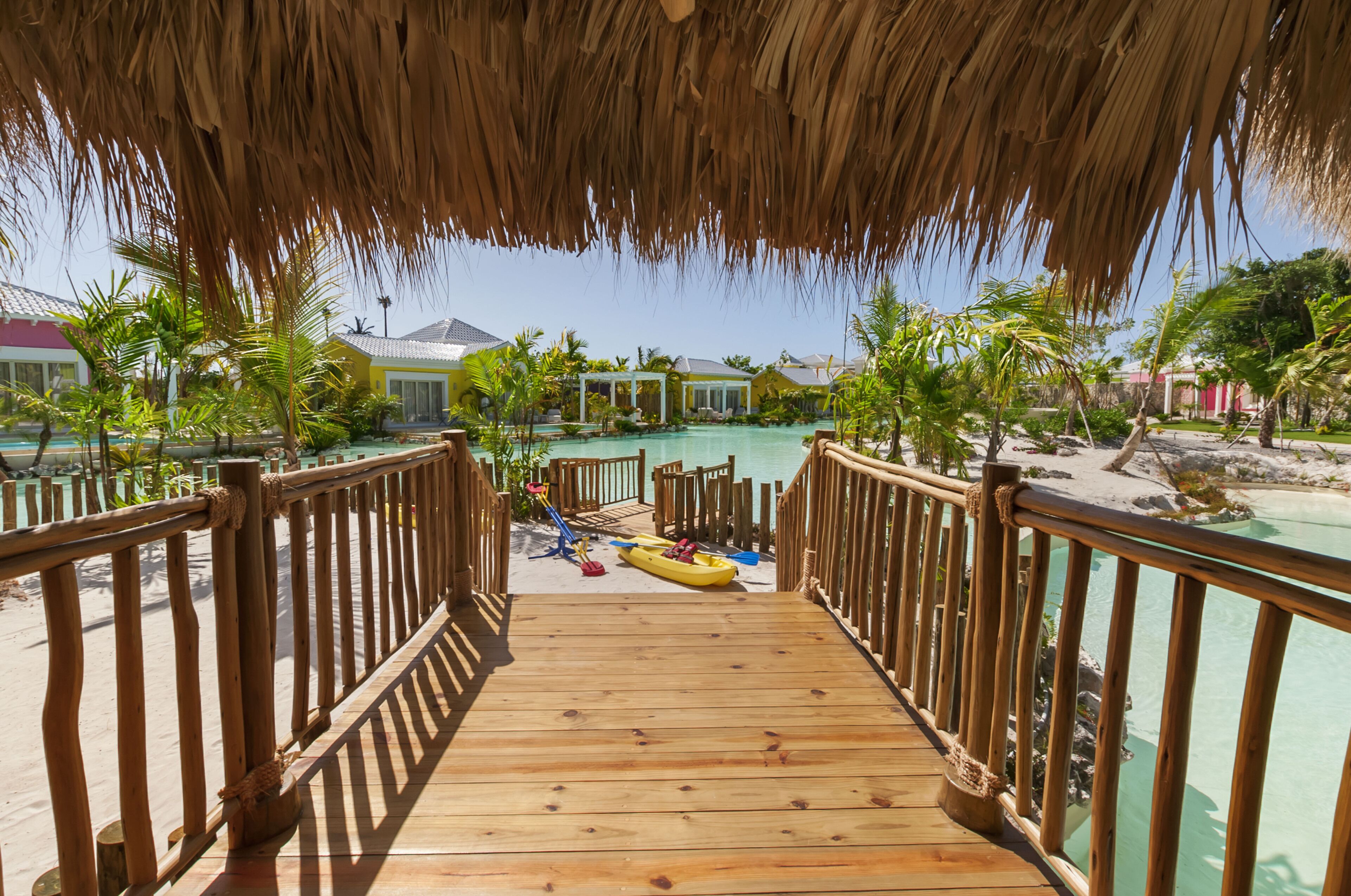 Une terrasse en bois mène à un espace piscine doté d’un toit de chaume, entouré de palmiers et offrant une vue dégagée sur l’océan.