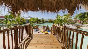 Une terrasse en bois mène à un espace piscine doté d’un toit de chaume, entouré de palmiers et offrant une vue dégagée sur l’océan.