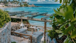 A wooden deck with seating and a table, overlooking a turquoise sea with palm trees and a thatched-roof structure in the background.