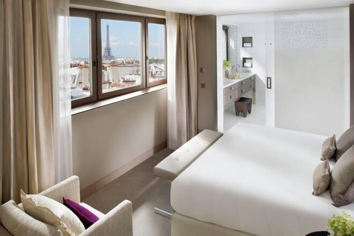 A hotel room with a bed, a chair, a desk, and a view of the Eiffel Tower.