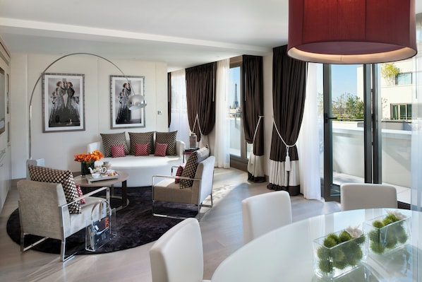A modern hotel room with a sofa, armchairs, a dining table, and large windows with curtains.