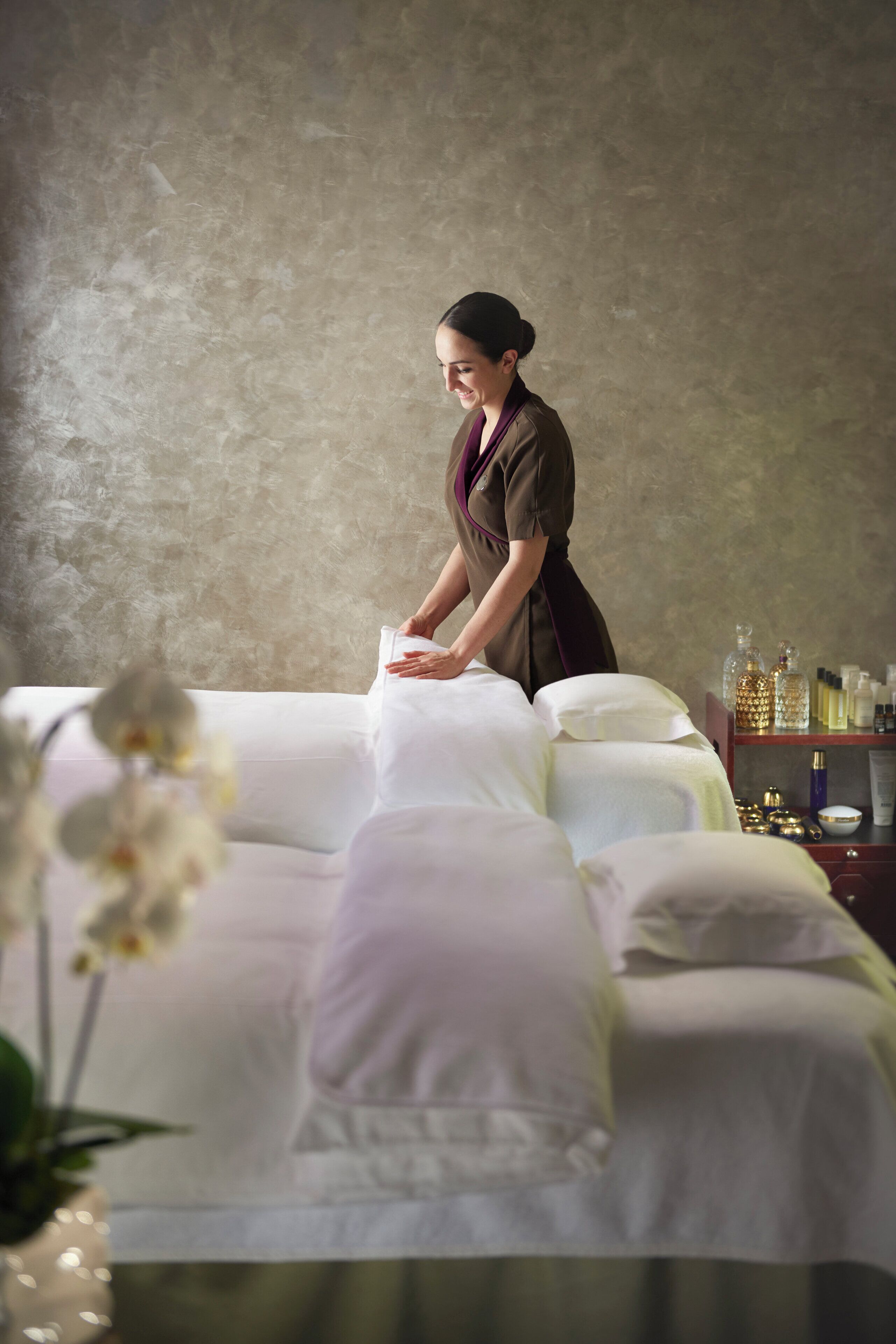 A spa room with a massage table, a massage therapist, and a shelf with various spa products.