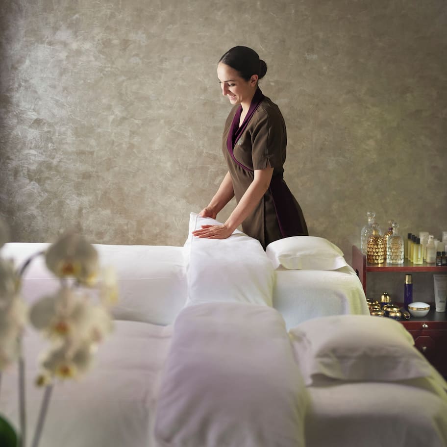 A spa room with a massage table, a massage therapist, and a shelf with various spa products.