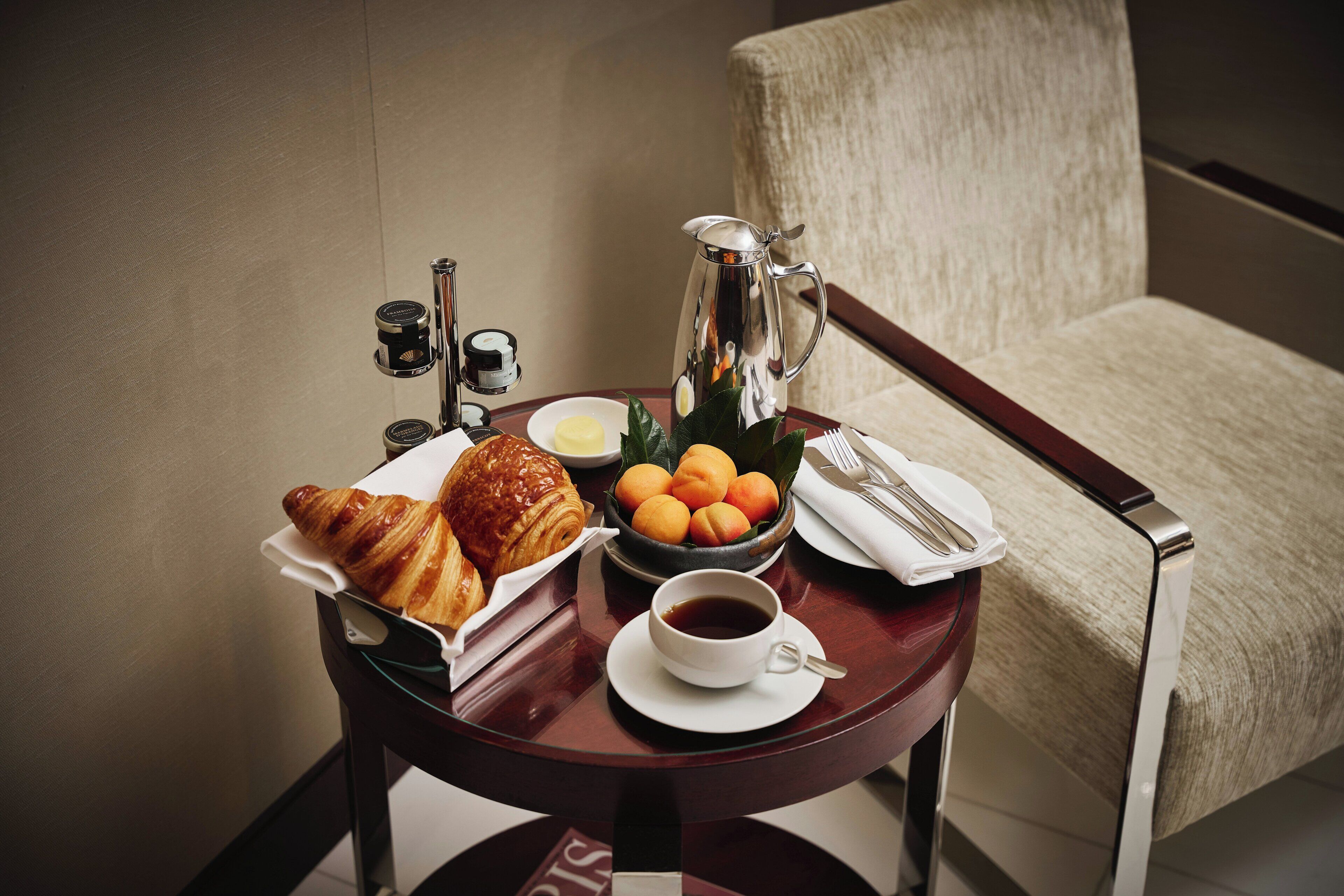 Ein Tisch mit Croissants, Orangen, einer Kaffeetasse und einem Teekessel vor einer beige farbenen Wand mit einem Stuhl.