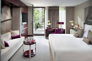 A hotel room with a bed, a sofa, a desk, and a small balcony with plants.