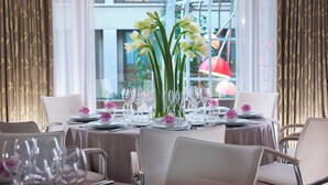 A dining table set with multiple chairs, a vase with flowers, and a view of a building through a large window.